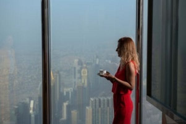 빨간 옷을 입은 사람이 컵을 들고 창가에 서서 흐릿한 하늘 아래 높은 건물이 있는 도시 풍경을 내려다보고 있습니다.