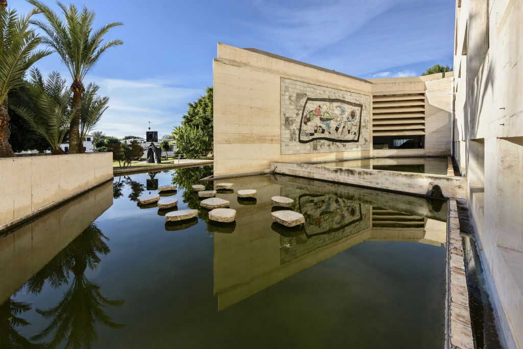 디딤돌, 나무, 맑고 푸른 하늘로 둘러싸인 수영장에 벽화가 반사된 대형 베이지색 건물.