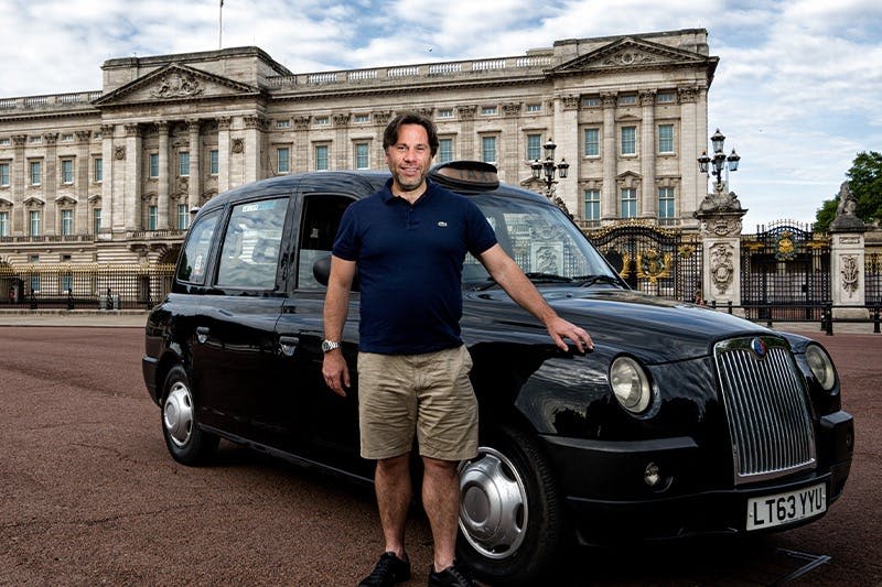 화려한 건축물이 있는 웅장한 건물 앞에 남색 셔츠와 카키색 반바지를 입은 남성이 검은색 택시 옆에 서 있습니다.