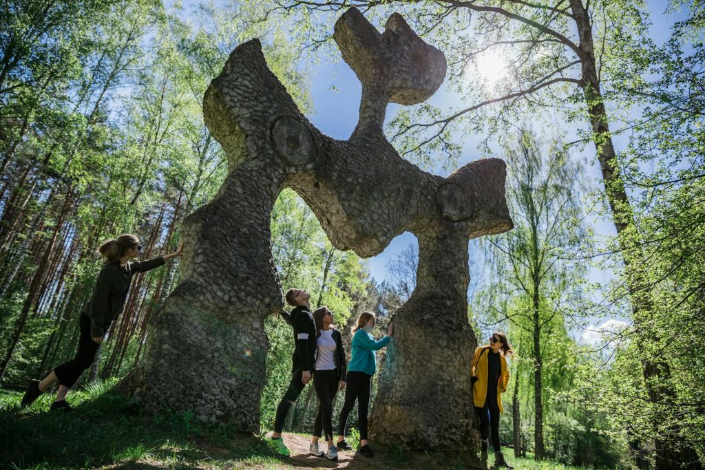 햇살이 내리쬐는 숲의 크고 추상적인 석조 조형물 아래 네 사람이 서 있습니다.
