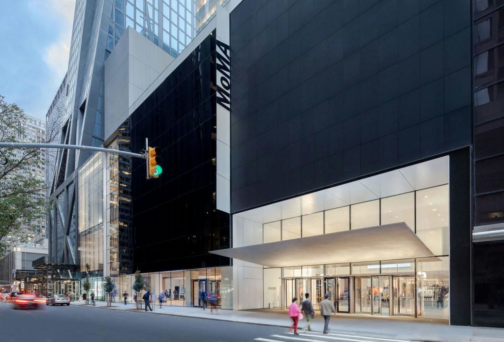 사람들은 외관에 "MoMA" 가 있는 현대적인 건물을 지나갑니다. 신호등과 횡단보도가 보입니다.