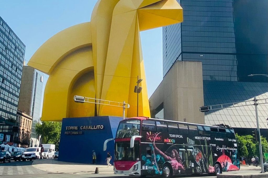 커다란 노란색 "Torre Caballito" 조각상을 배경으로 한 2층 투어 버스가 있습니다.