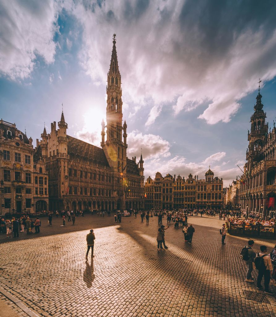 역사적인 건물, 높은 중앙 타워, 걷고 모이는 사람들이 있는 햇살이 내리쬐는 광장. 하늘은 부분적으로 흐립니다.