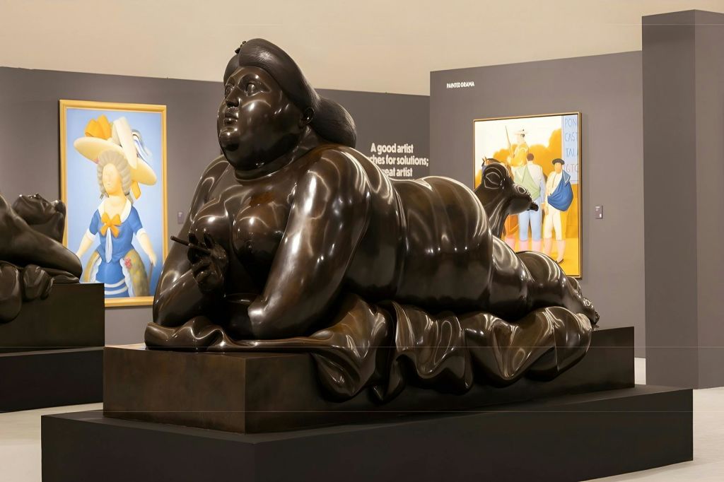 갤러리에 전시된 리클라이닝 여성의 청동 조각품으로, 그림이 배경에 보입니다.