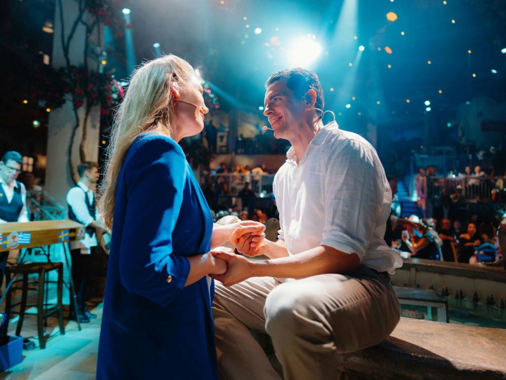 무대에서 한 남성과 한 여성이 생동감 넘치는 관객과 화려한 조명을 배경으로 대화를 나누고 있습니다.