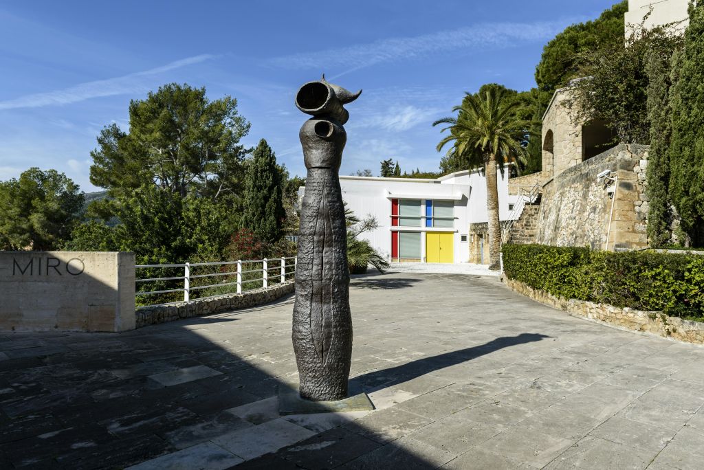 원색 창문을 배경으로 현대적인 흰색 건물이 있는 마당에 높이 솟은 추상적인 조형물이 있습니다.