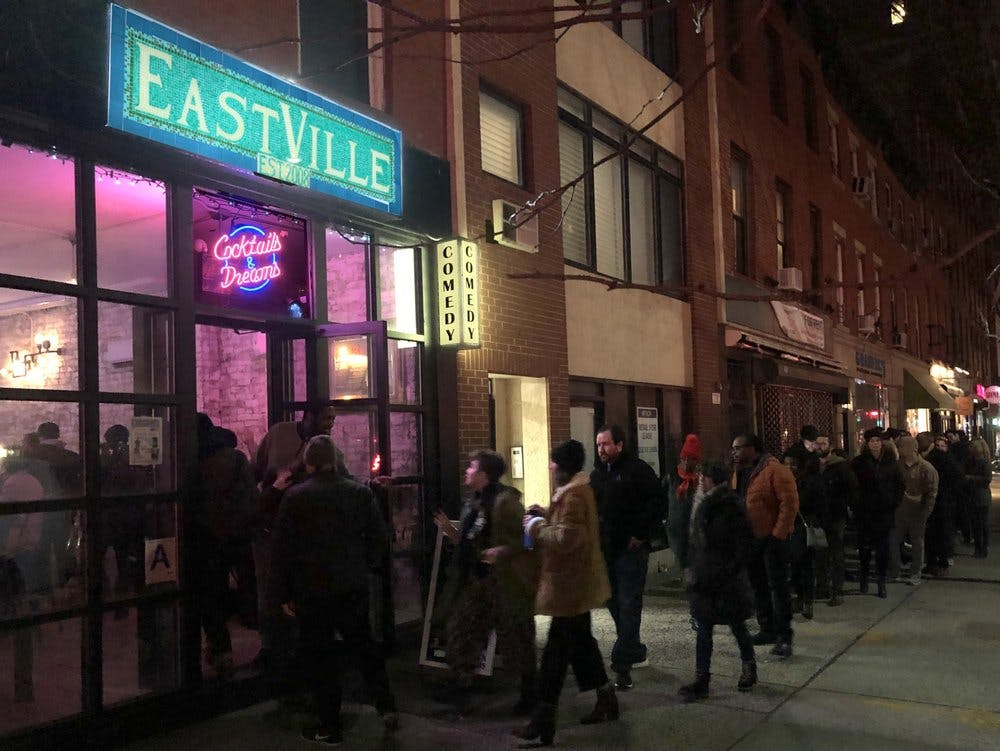 밤에 도시의 거리에서 코미디 클럽 밖에서 한 줄의 사람들이 기다리고 있습니다. 네온 사인에는 "웨스트빌" 이라고 쓰여 있습니다.