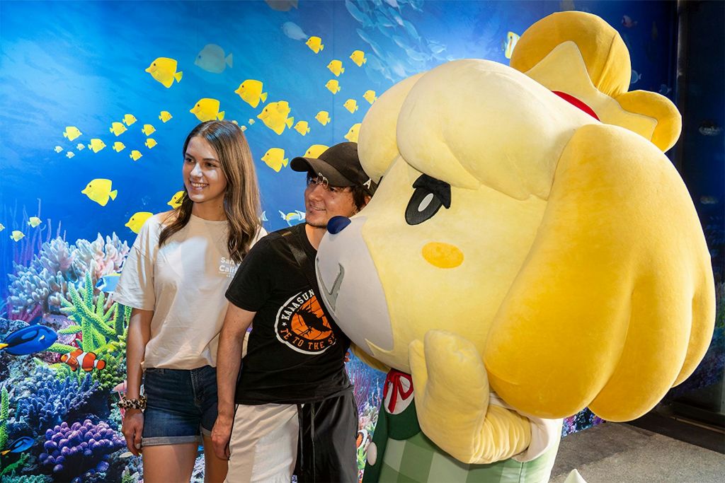 물고기와 산호를 배경으로 수중을 테마로 한 커다란 봉제인형 옆에 서 있는 두 사람.