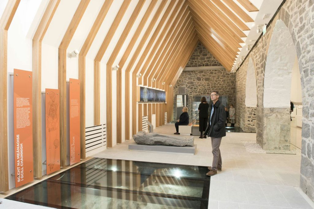 돌담, 나무 대들보, 투명한 바닥 부분, 바위 조각품이 전시된 현대적인 갤러리의 방문객들.