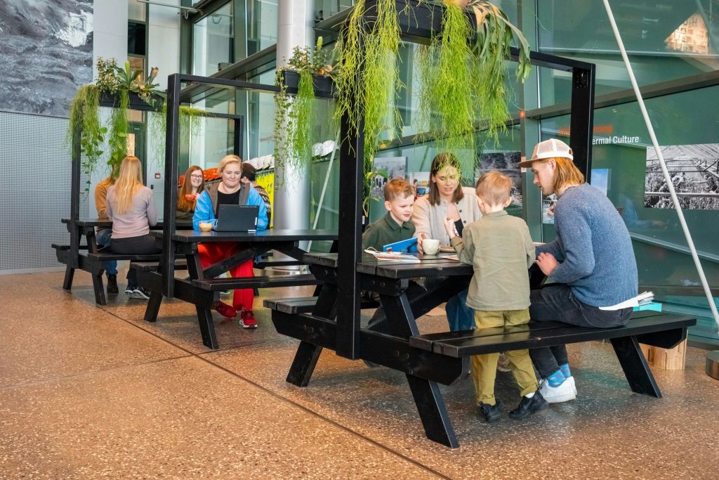 실내 분위기의 피크닉 스타일 테이블에 앉아 있는 사람들로, 머리 위에는 식물이 걸려 있고 모던하고 밝은 인테리어 디자인이 돋보입니다.