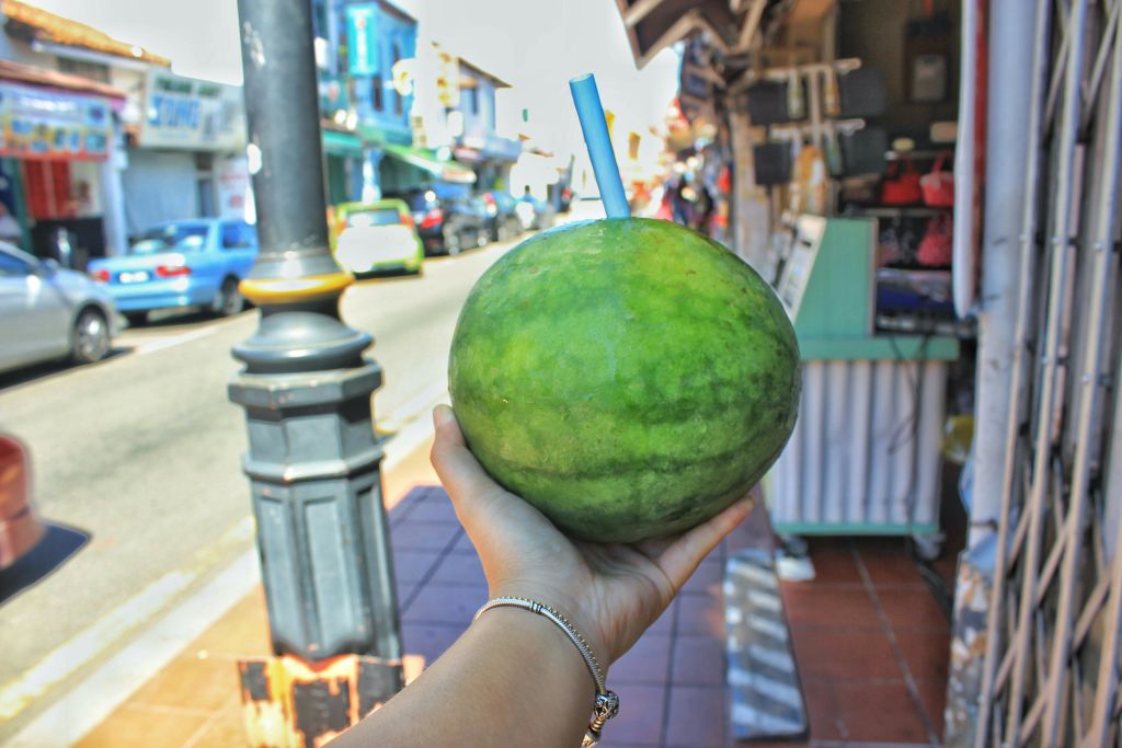 파란색 빨대가 삽입된 수박을 통째로 들고 거리와 상점을 배경으로 손을 잡습니다.