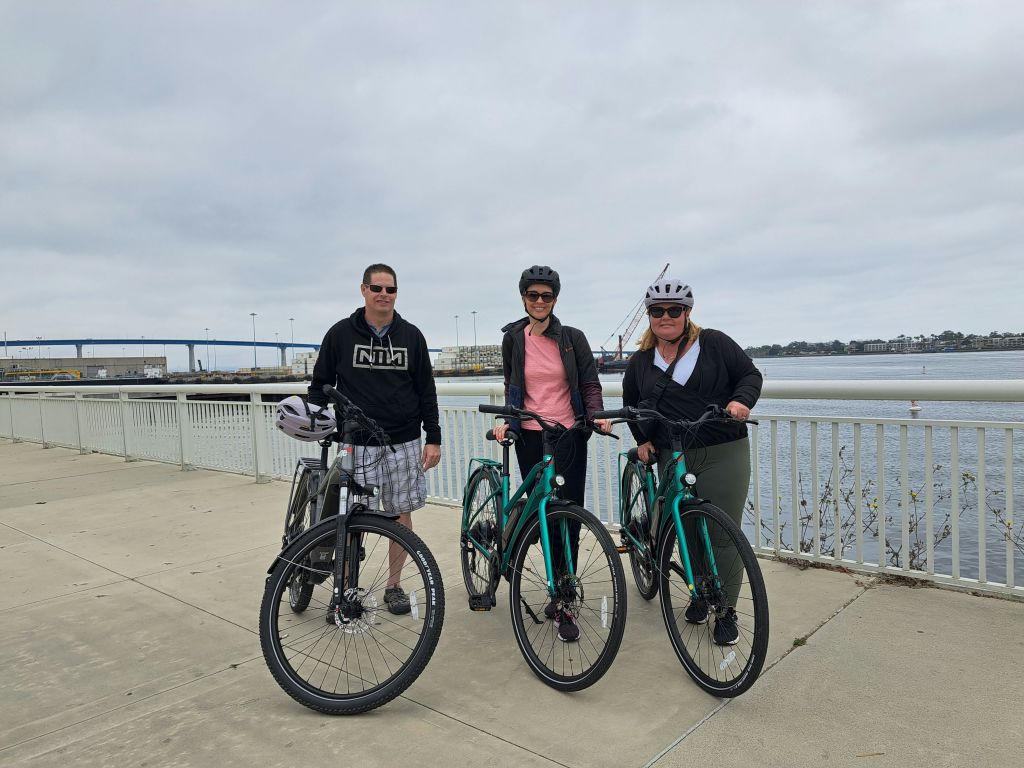 수변 산책로에 자전거를 탄 세 사람. 그들은 헬멧과 캐주얼한 옷을 입고 있습니다. 배경에는 다리가 있습니다.