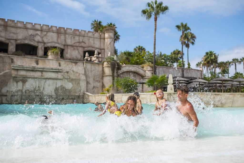 성곽 같은 구조와 야자수를 배경으로 얕은 수영장에서 물놀이를 즐기는 가족.