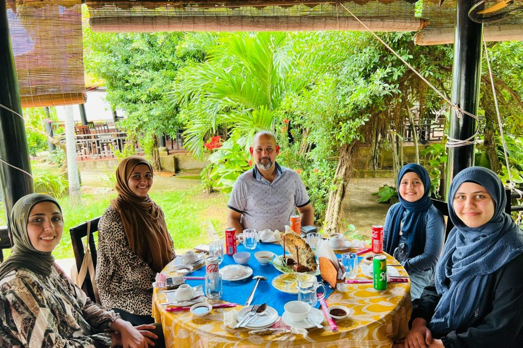 푸르른 녹음으로 둘러싸인 야외 테이블에 세 사람이 앉아 음식과 음료를 즐기고 있습니다. 식탁보가 노란색입니다.