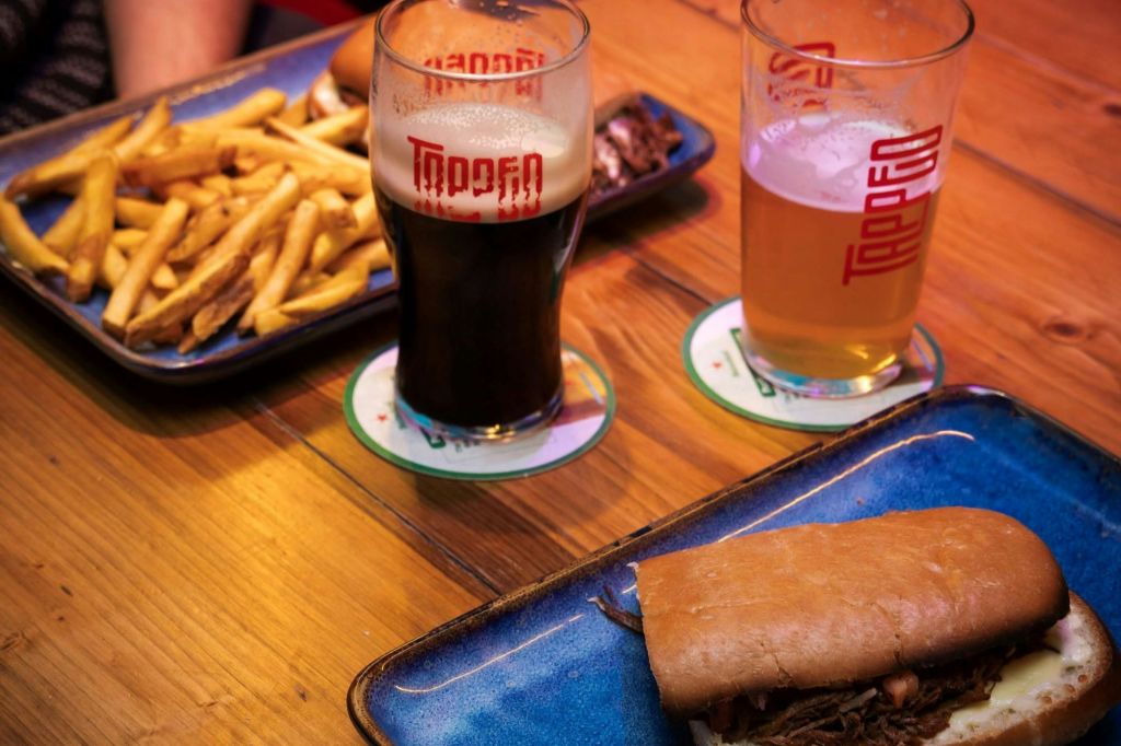 맥주 두 잔, 감자튀김이 담긴 접시, 나무 테이블 위에 놓인 샌드위치, 파란색 접시 위에 브랜드 컵받침이 놓여 있습니다.