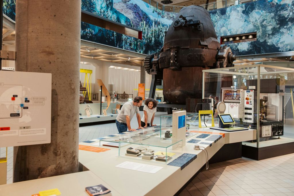 두 사람이 대형 산업 기계와 유리 케이스 아래 다양한 디스플레이가 있는 박물관의 전시물을 살펴보고 있습니다.