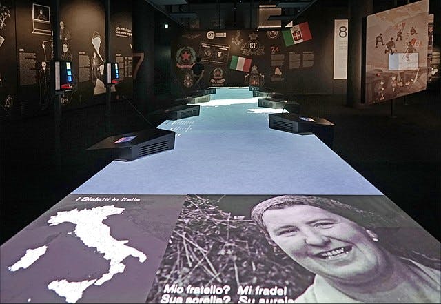 역사적 이미지, 이탈리아 지도, 이탈리아어로 된 텍스트를 담은 인터랙티브 전시. 플래그 디스플레이 및 프로젝션 화면이 표시됩니다.