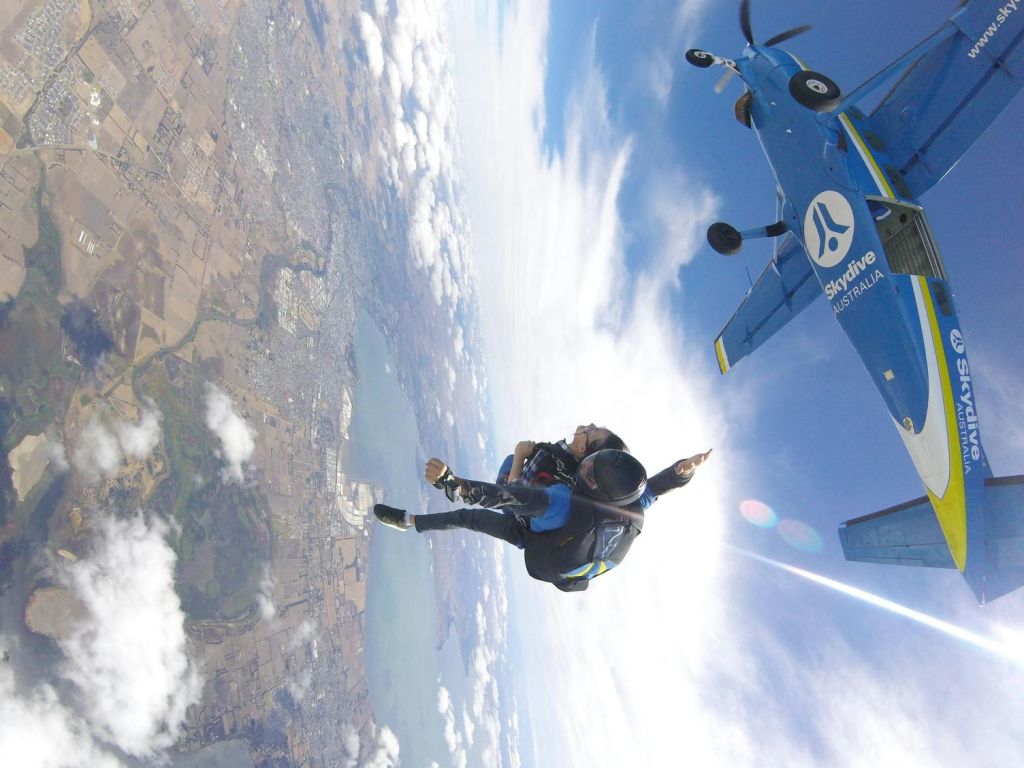 "스카이다이브 오스트레일리아 (Skydive Australia)" 비행기를 배경으로 두 사람이 탠덤 스카이다이빙을 즐기고 있습니다. 밭과 물이 흐르는 풍경 위로 높이 솟아 있습니다.
