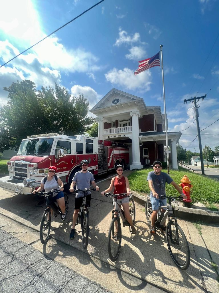 맑은 하늘 아래 바깥에 소방차가 주차된 소방서 앞에서 자전거를 탄 네 명의 사람들.