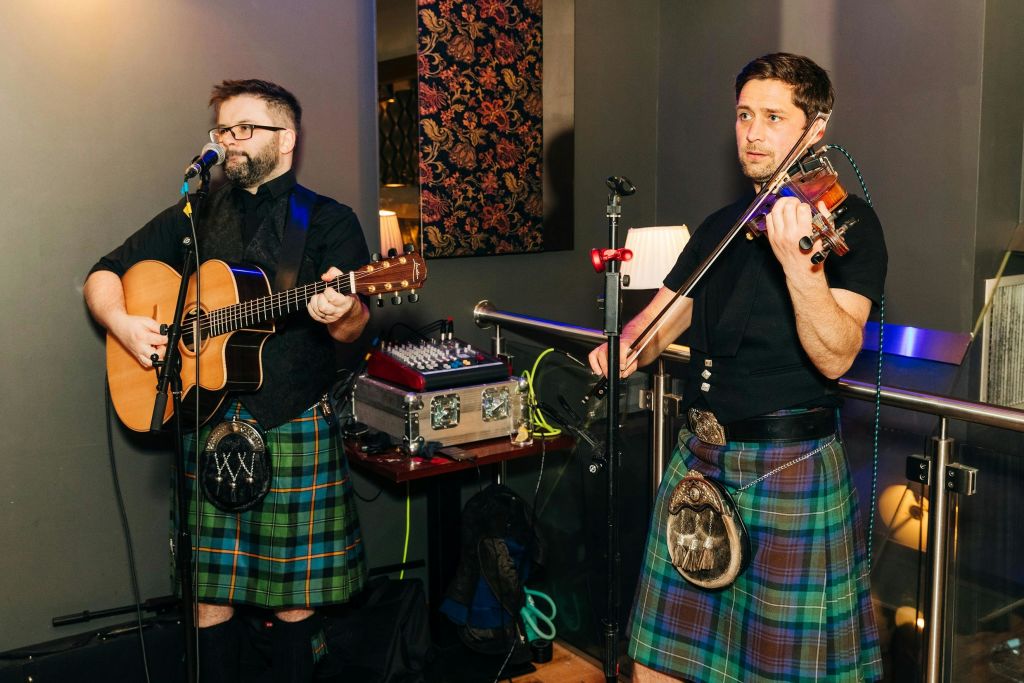 킬트를 입은 남성이 실내에 서서 바이올린을 연주하고 있으며, 뒤에는 음향 장비가 보입니다.