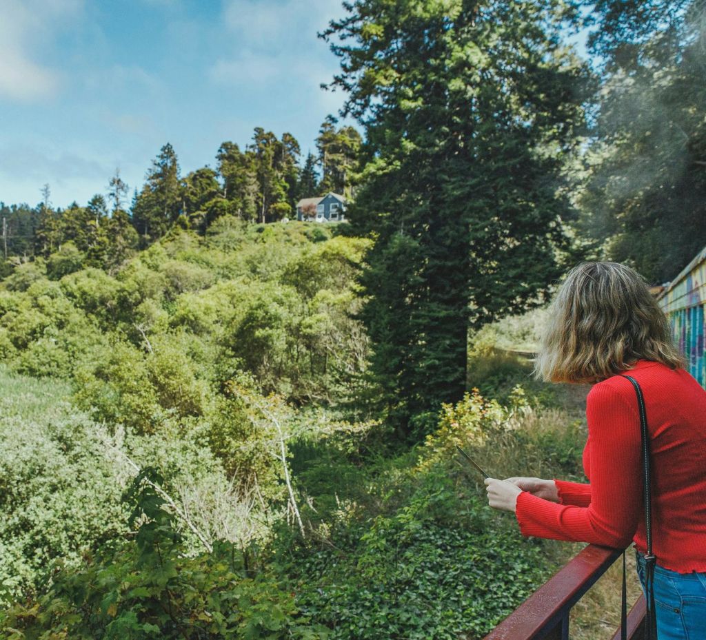 빨간 스웨터를 입은 사람이 발코니에 서서 나무가 우거진 푸른 언덕과 저 멀리 보이는 집을 내려다보고 있습니다.