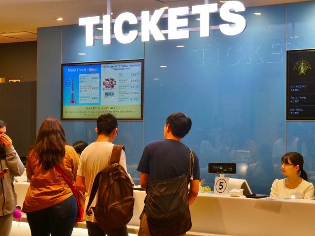 조명이 켜진 커다란 'TICKETS' 표지판 아래 점원이 있는 티켓 카운터에 세 사람이 서 있습니다. 디지털 화면에는 가격이 표시됩니다.