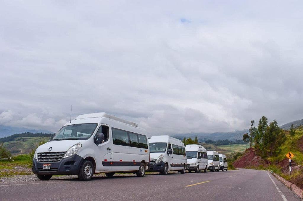 잔디밭과 산을 배경으로 흐린 하늘 아래 시골길에 흰색 승합차가 줄지어 주차되어 있습니다.