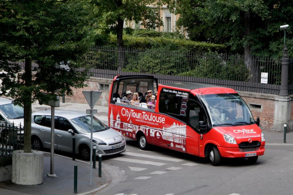 "시티 투어 툴루즈" 라고 적힌 빨간색 오픈탑 투어 버스에 사람들이 앉아 있습니다. 나무가 늘어선 거리의 은색 자동차 옆에 있습니다.