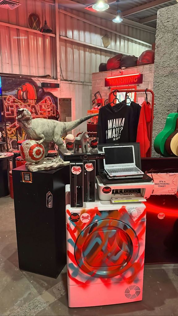 네온사인, 공룡 모형, 노트북, 다양한 물품이 있는 방. "Wanna Smash?" 라고 적힌 셔츠와 벽에 "Warning" 표시가 있습니다.