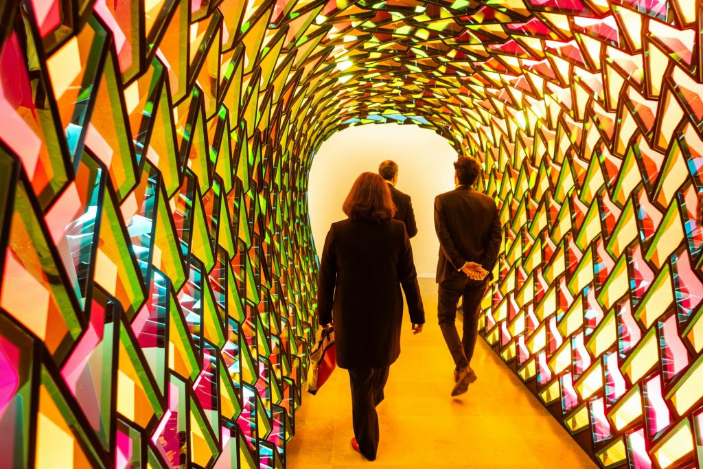 세 사람이 기하학적인 스테인드글라스 패널로 만든 다채롭고 빛나는 터널을 거닐고 있습니다.