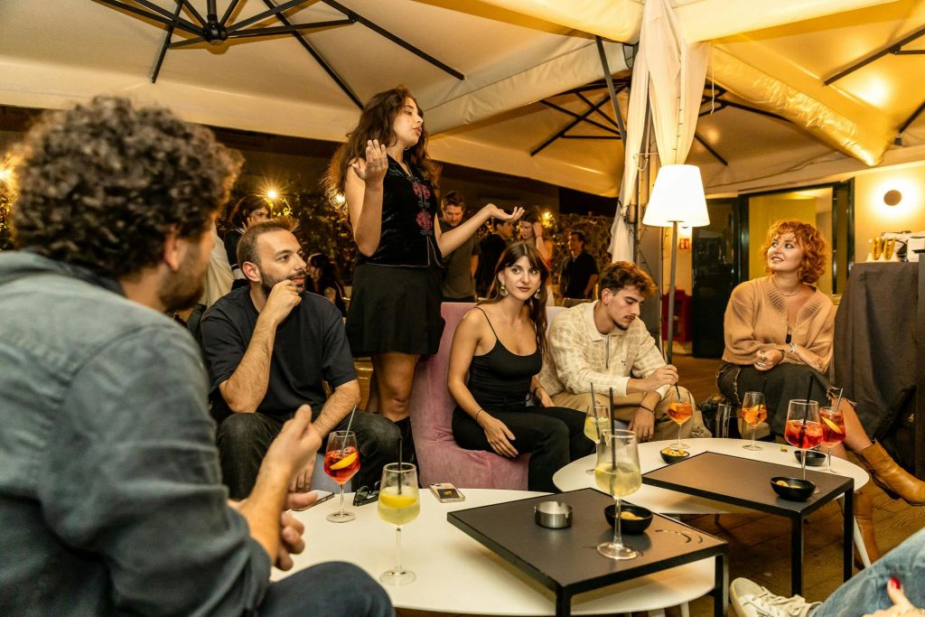 캐노피 아래에서 음료를 마시며 테이블에서 사람들과 어울리는 한 무리의 사람들, 한 사람이 서서 열정적으로 제스처를 취합니다.