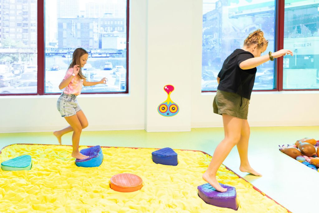 도시를 배경으로 커다란 창문이 있는 노란색 패딩 방에서 화려한 쿠션 위에서 노는 소녀.