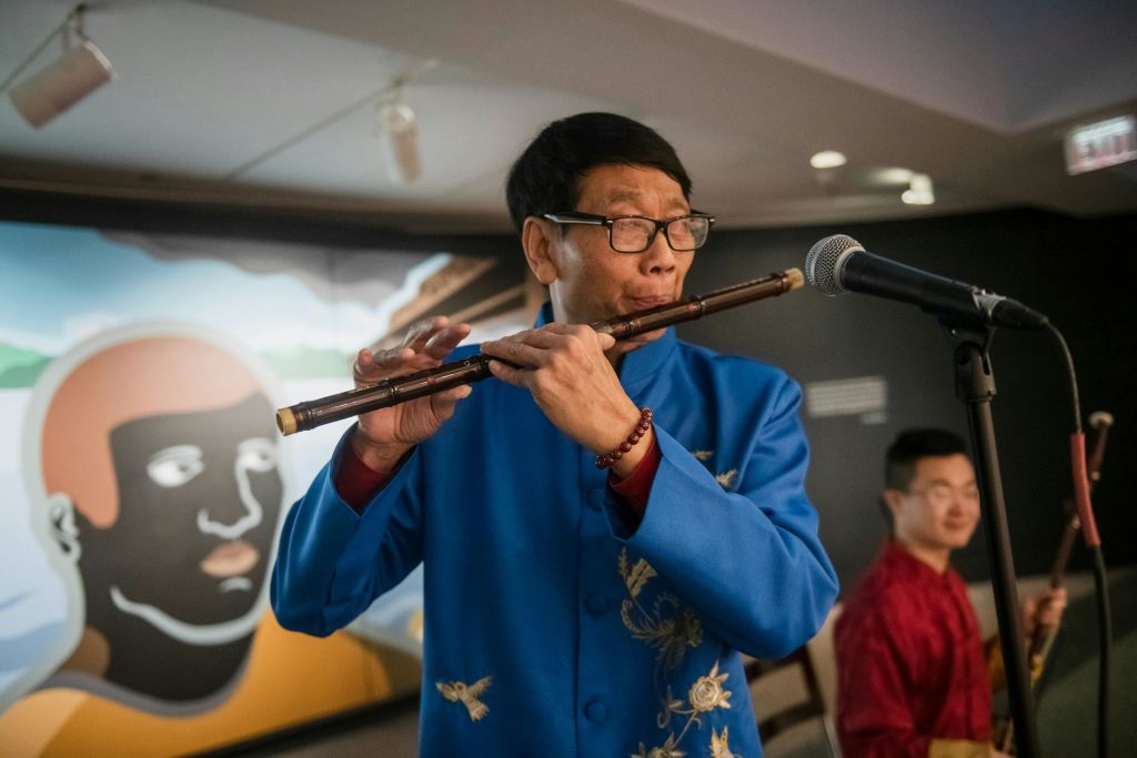 얼굴 벽화를 배경으로 플루트를 연주하는 파란색 자수 재킷을 입은 남성.