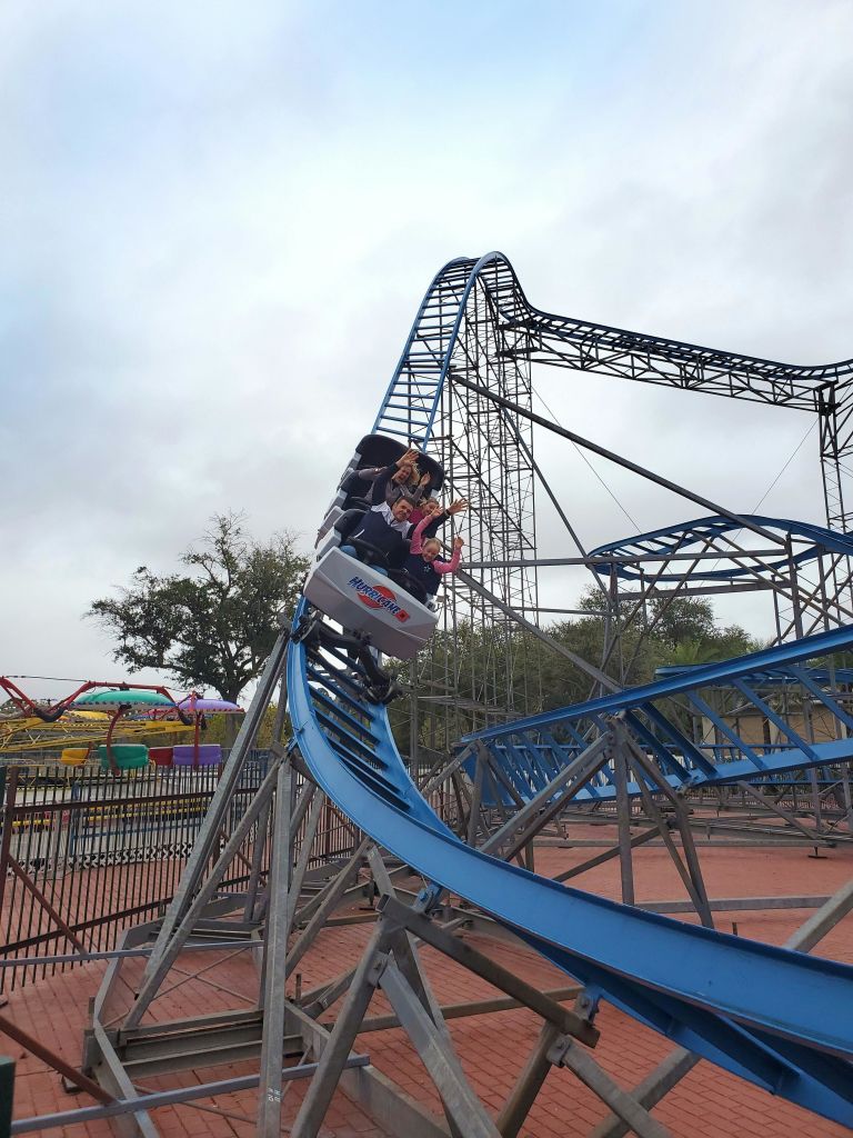 놀이공원에서 파란색 금속 롤러코스터를 타는 사람들, 다채로운 놀이기구와 나무가 배경입니다.