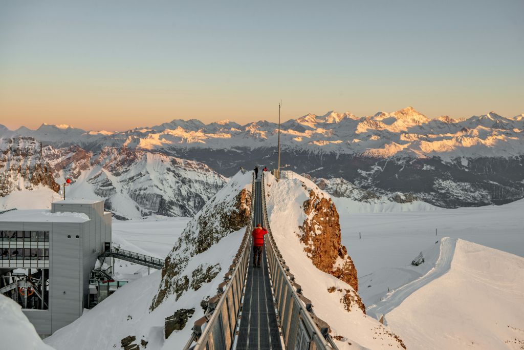 붉은 재킷을 입은 사람이 해가 지는 동안 눈 덮인 산을 가로지르는 좁은 다리를 걸으며 먼 봉우리를 비춥니다.