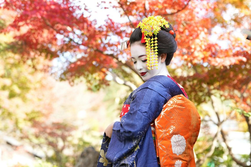 하얀 얼굴 메이크업을 한 일본 전통 복장을 한 사람이 활기찬 가을 단풍 사이에 서 있습니다.