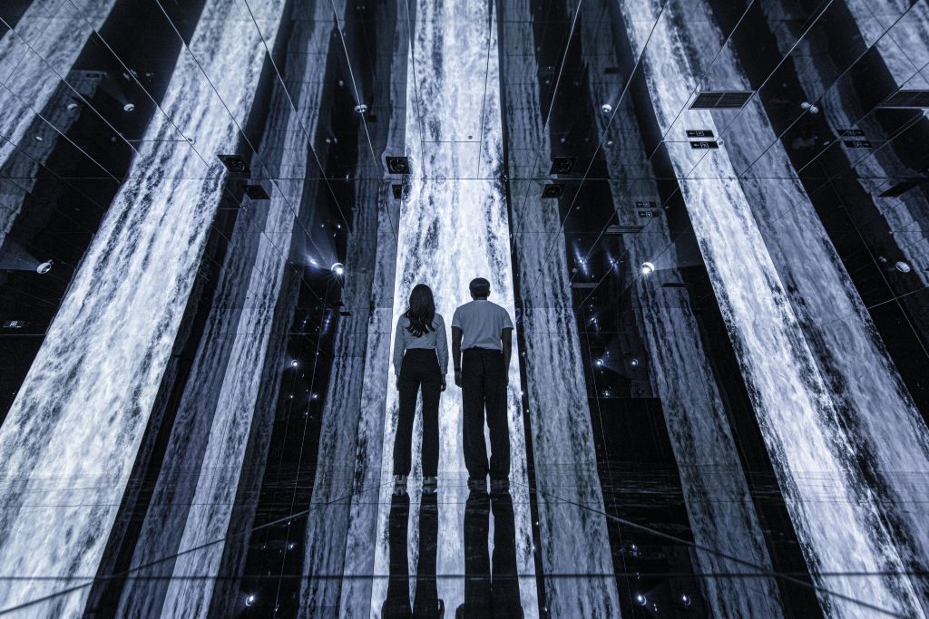 조명이 켜진 수직 조명 스트립이 있는 거울 방에 서 있는 두 사람은 미래 지향적이고 반사적인 환경을 조성합니다.