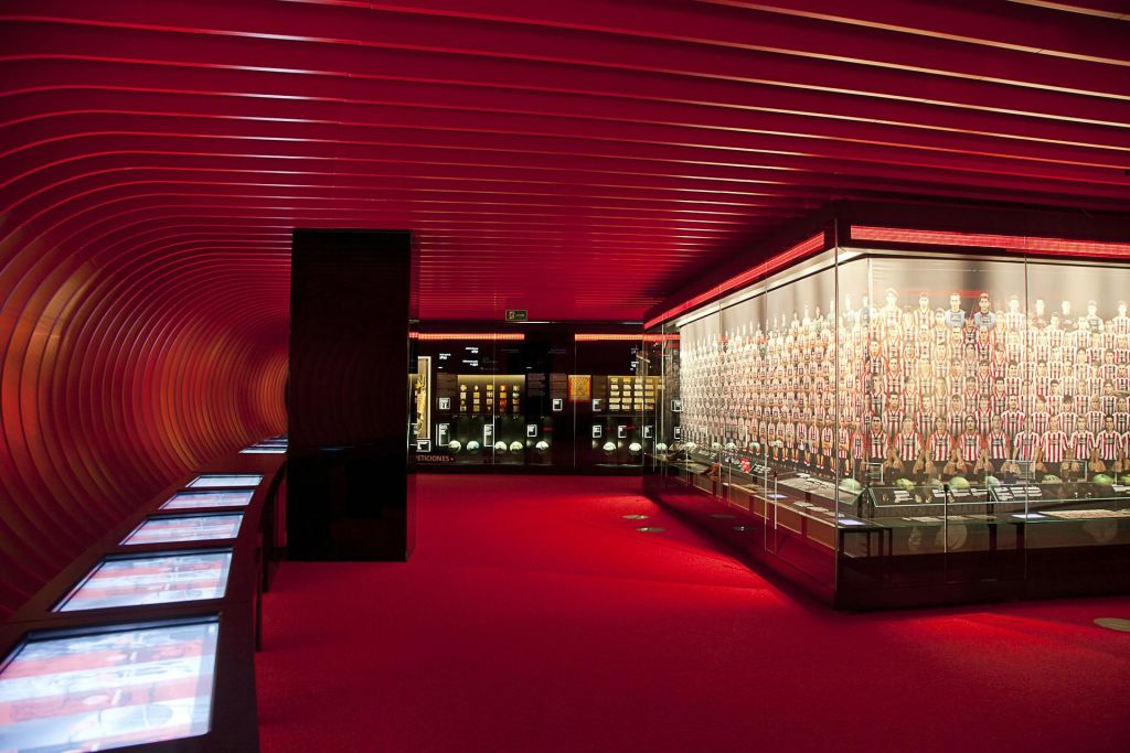 빨간색 벽과 카펫이 있는 객실로, 디지털 정보 화면이 있는 조명 유리 디스플레이에 스포츠 기념품을 전시하고 있습니다.