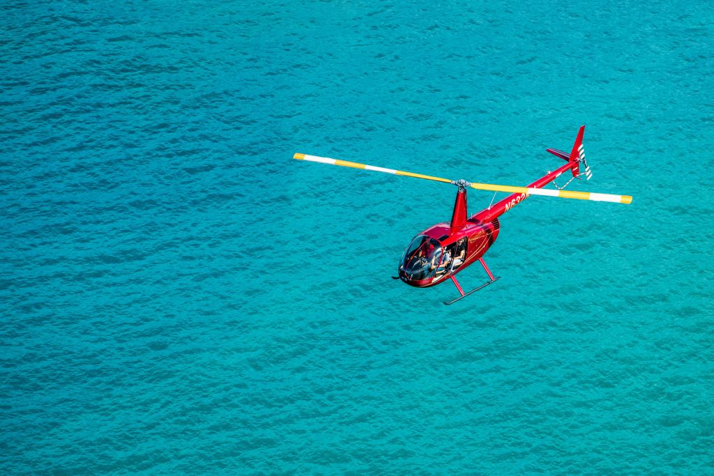 노란색 끝이 있는 로터 블레이드가 있는 빨간색 헬리콥터가 청록색의 맑은 물 위로 날아갑니다.