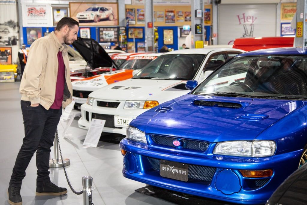 베이지색 재킷을 입은 한 남성이 자동차 박물관 내부에서 파란색 스포츠카를 바라보고 있으며, 다른 자동차와 자동차 포스터를 배경으로 하고 있습니다.