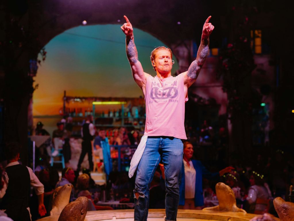 분홍색 셔츠와 청바지를 입은 사람이 활기찬 레스토랑 분위기에 둘러싸인 채 팔을 들어 올린 채 무대에 서 있습니다.
