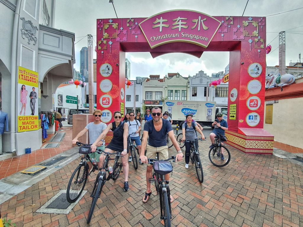 '차이나타운 싱가포르' 를 배경으로 빨간색 아치형 통로 아래에서 자전거를 탄 사람들이 포즈를 취하고 있습니다.