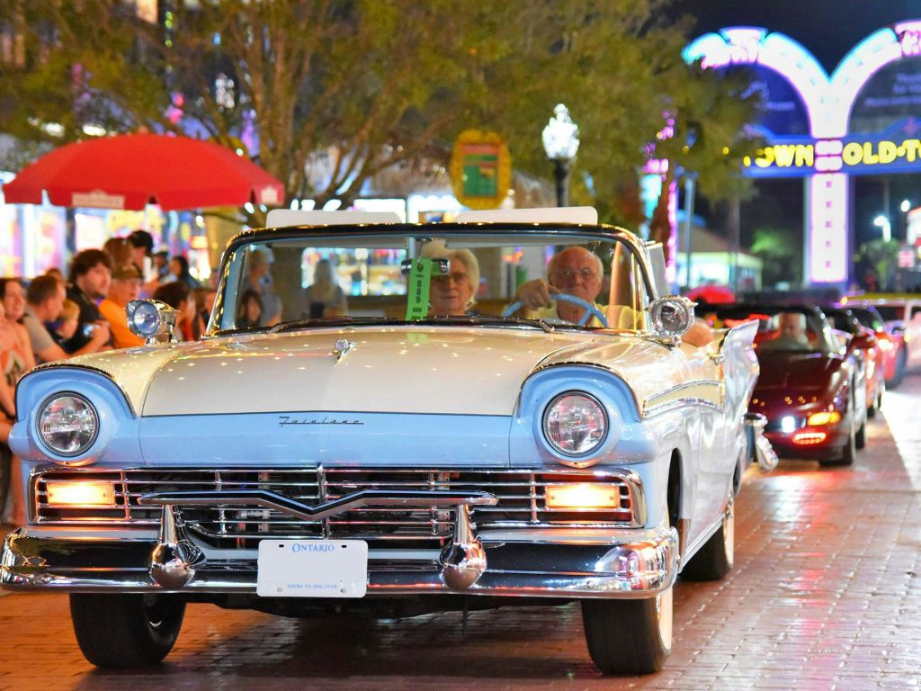 빈티지한 파란색과 흰색의 컨버터블 자동차가 밤에는 축제 분위기가 물씬 풍기는 거리를 배경으로 운전합니다.