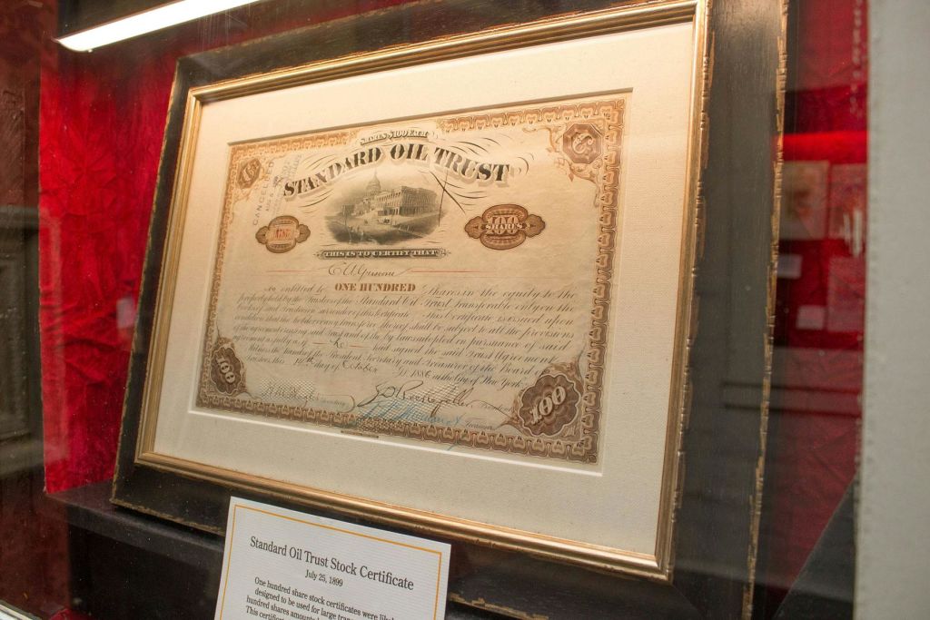 액자에 담긴 스탠다드 오일 트러스트 (Standard Oil Trust) 주식 증서는 아래에 설명이 있는 명판이 있는 벽에 표시됩니다.