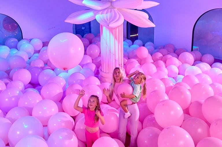 커다란 꽃 장식 아래 분홍색 풍선이 가득한 방에 한 여성과 두 자녀가 서 있습니다.