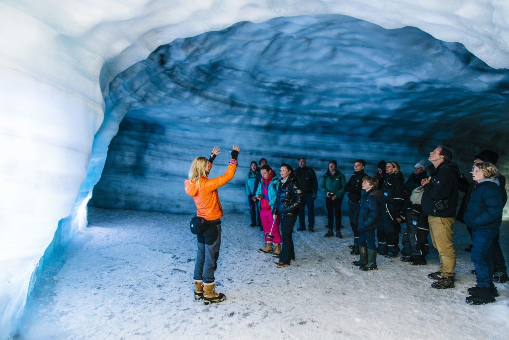 겨울 옷을 입은 한 무리의 사람들이 질감이 살아있는 푸른 벽과 눈 덮인 땅이 있는 얼음 동굴 내부에서 가이드의 말을 듣고 있습니다.
