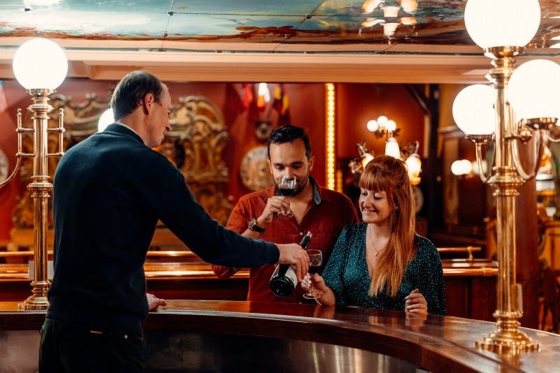 바에 있는 세 사람: 한 남자와 한 여자를 위해 와인을 붓는 바텐더. 배경에는 따뜻한 조명과 화려한 인테리어가 돋보입니다.