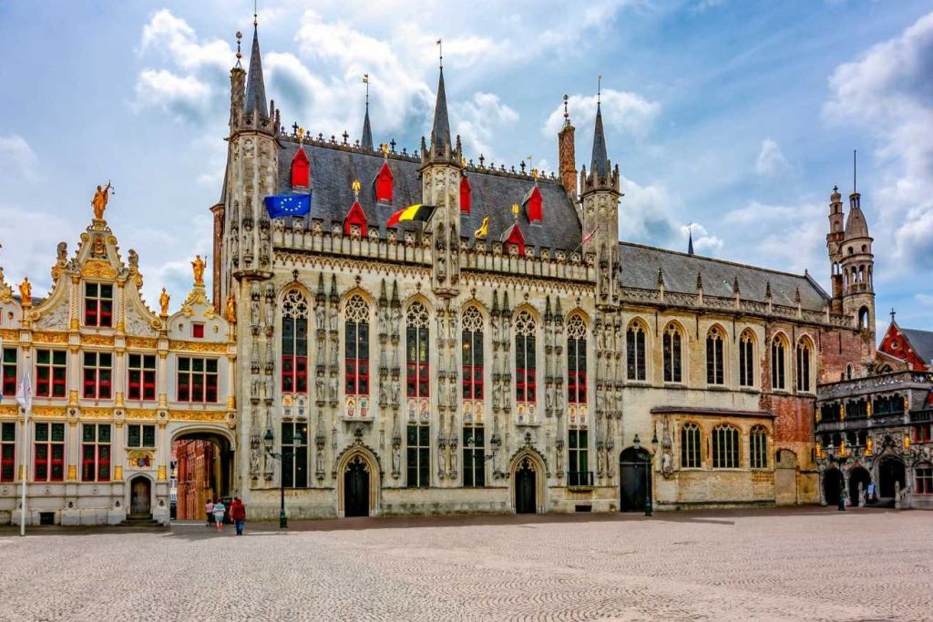 화려한 디테일, 첨탑, 깃발이 있는 고딕 양식의 건물. 인접한 아치형 통로. 전경의 자갈 광장. 왼쪽에 있는 사람들.