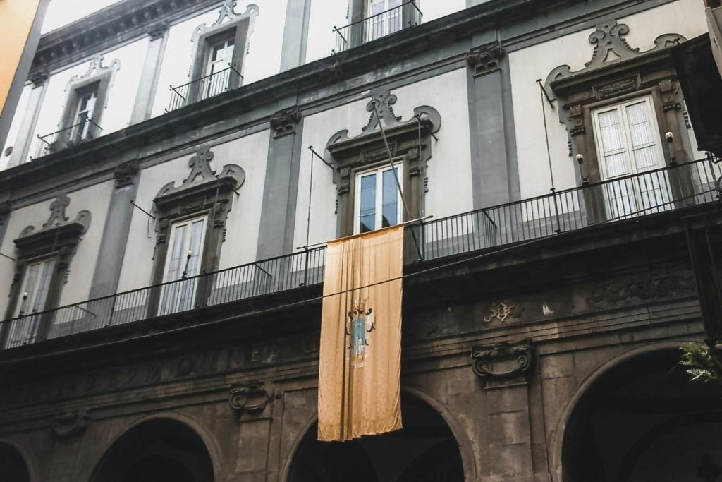 회색과 흰색 건물에 아치형 출입구와 발코니에 걸린 모습을 보여주는 노란색 배너가 있습니다.