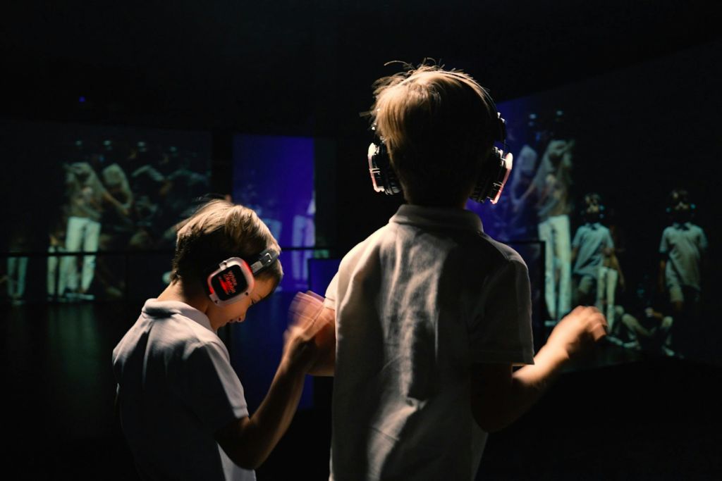 어두운 방에서 헤드폰을 착용한 두 어린이가 춤을 추고 있으며, 화려한 화면이 배경으로 사람들을 보여주고 있습니다.